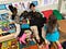 A smiling teacher sitting on a small couch under a pile of students who are hugging them.