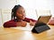 A young African American girl pays attention happily to her iPad tablet, learning remotely with a pencil in hand.
