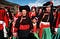 Mosuo women and girls, ranging from young kids to elderly women, wearing dresses with red, black, green, and white designs, holding hands outdoors.