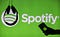 Headphones hang upside down as a hand holds a phone with the Spotify logo displayed in front of a screen showing Spotify