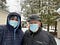 Father and son stand in front of the cabin while it’s snowing dressed in winter coats, hats, and surgical masks.