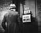 A black and white archival photo of a man standing in front of a door. The sign on the door says: ‘NO WORK. DON’T APPLY.’