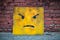Yellow box with an angry face against a brick wall.