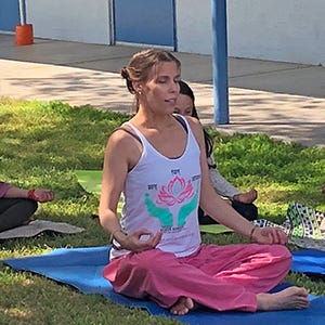 A woman sits meditating in Sukhasana, or easy-seated posture, on a yoga mat, on a grass lawn, surrounded by children.
