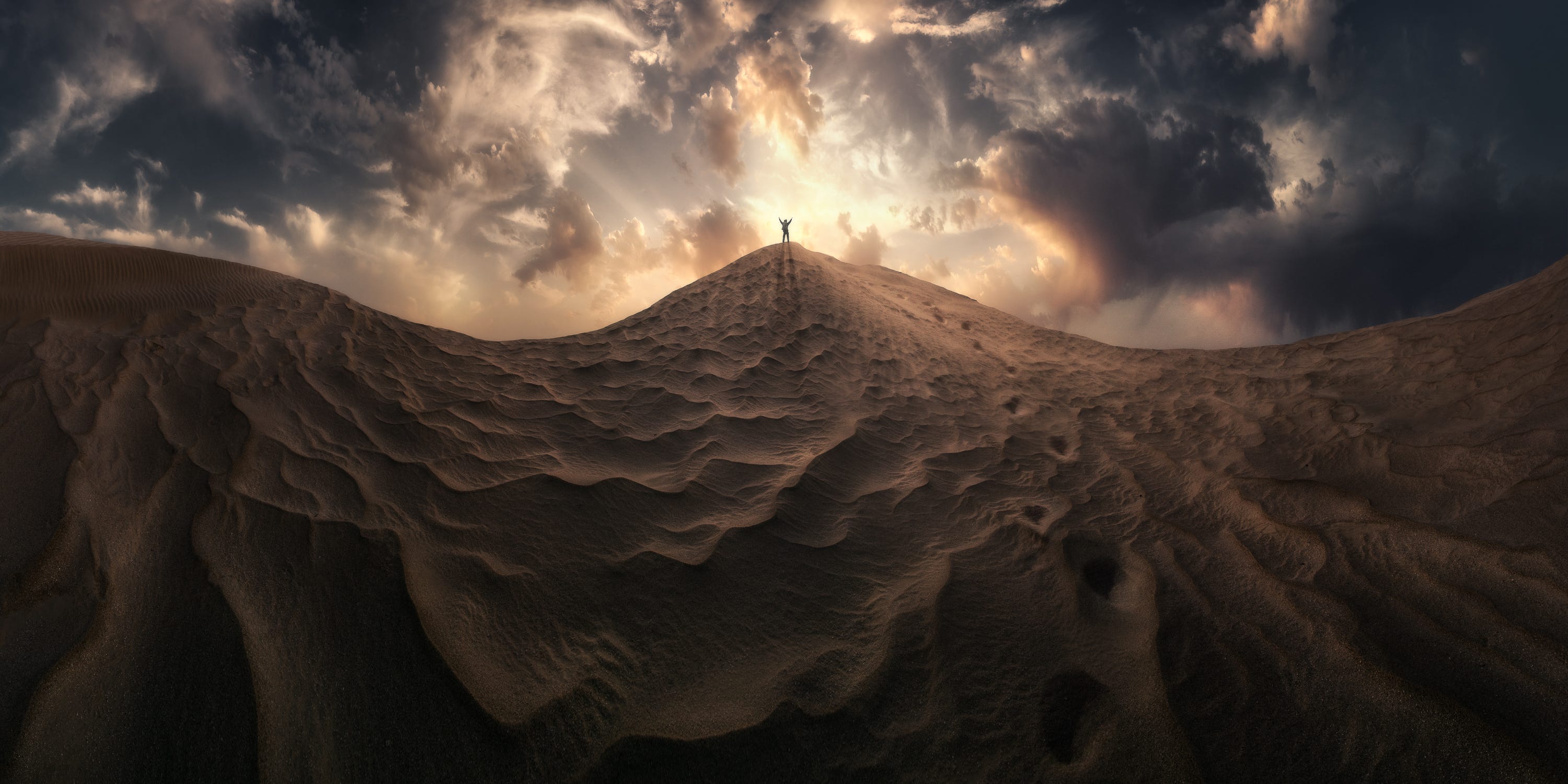 A man stands on top of peak of sand with arms outstretched, backed by a bright sun
