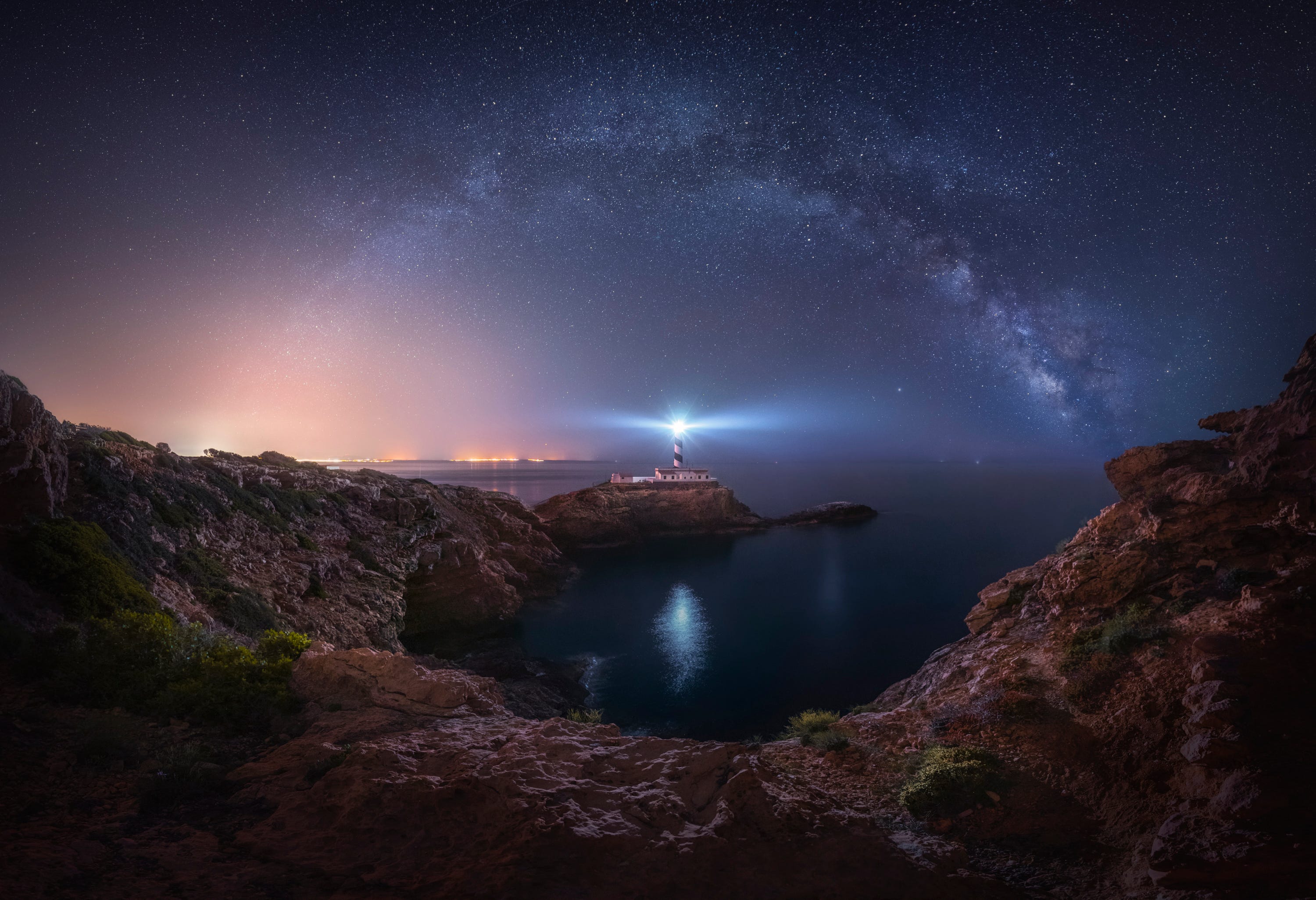 A small striped lighthouse shines a bright light over a rocky, dark ocean cove, with a very starry night sky shown above