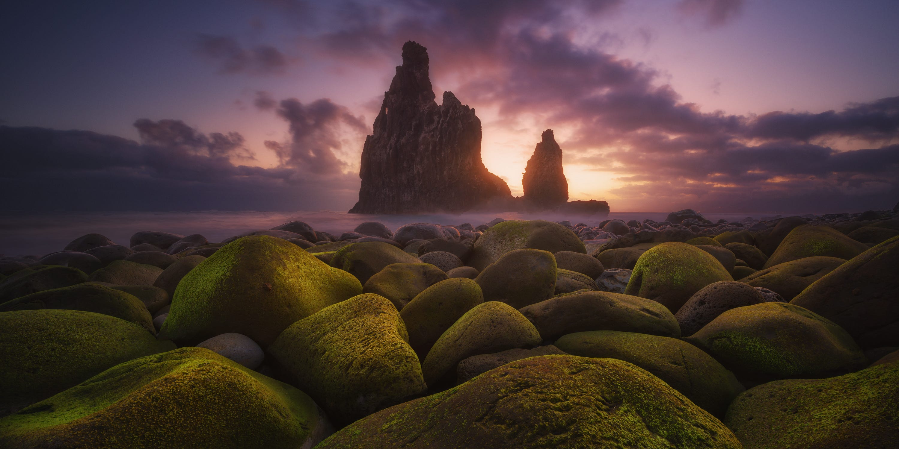 A fine art photograph of a craggy island backed by a purple and yellow sky and with mossy green rocks in the foreground