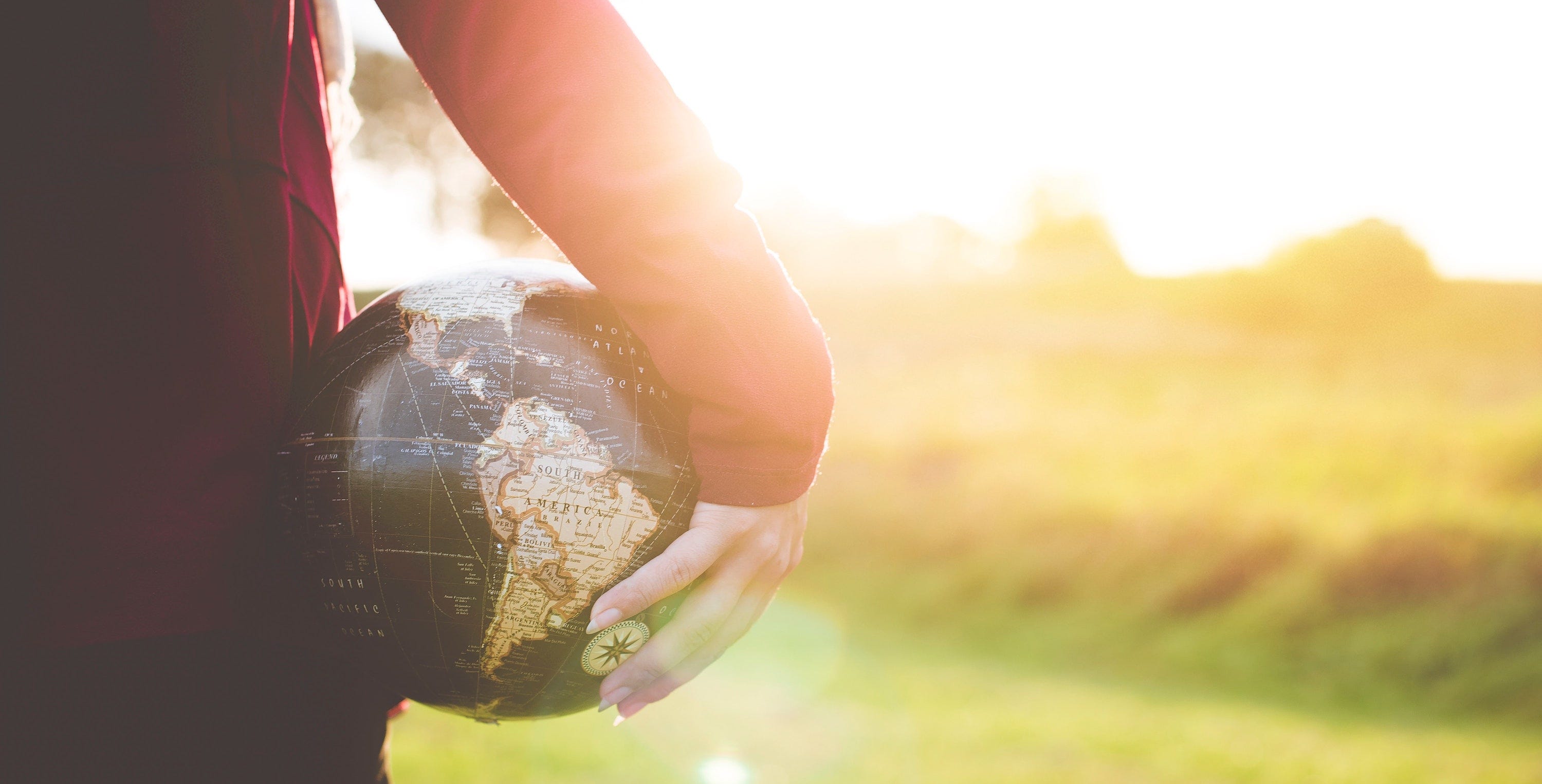 The back of a woman in a red shirt, holding a globe against her body with one hand as she looks out into bright sunlight