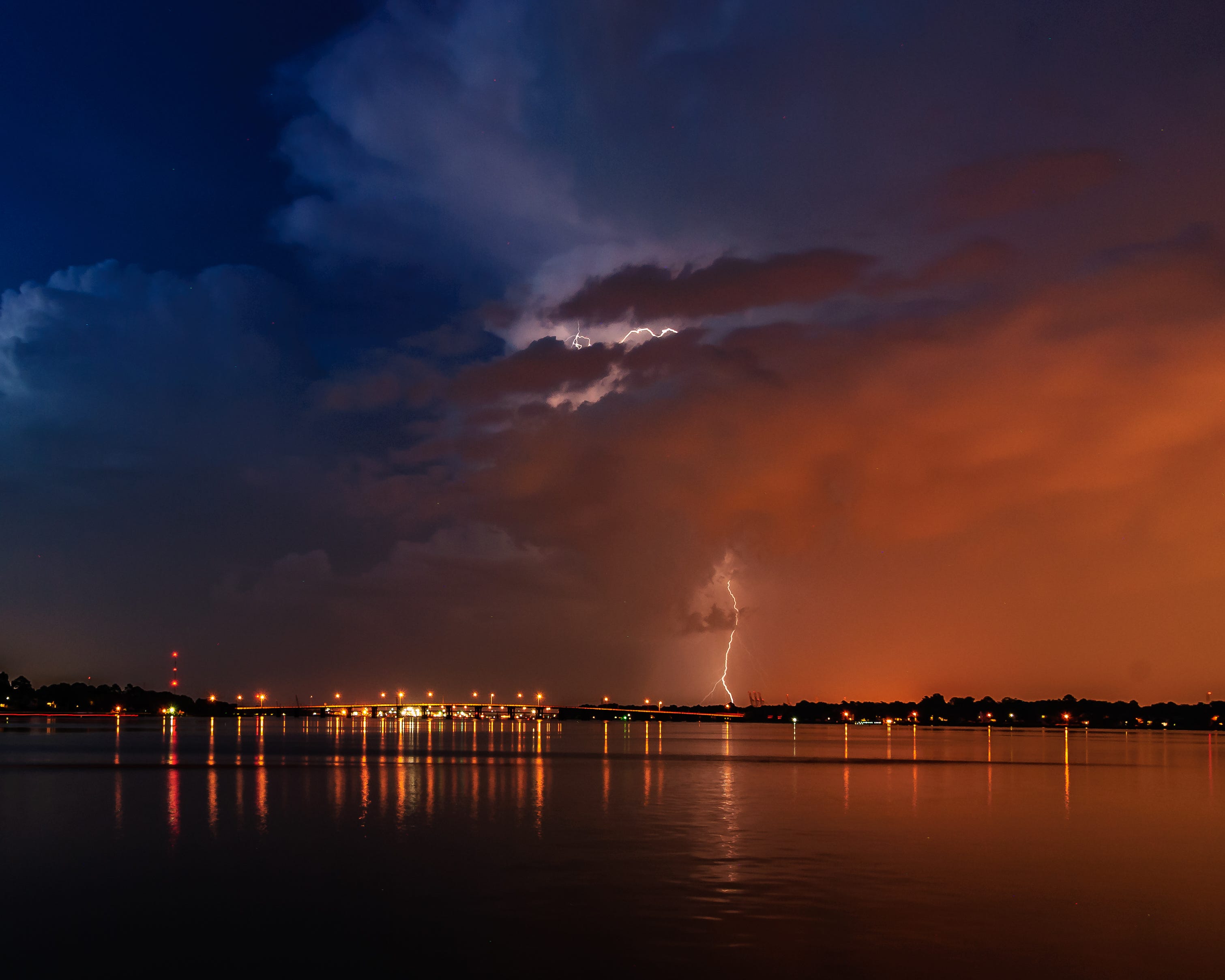 Lightening strikes light up the clouds above a body of water while lightening strikes the shore.