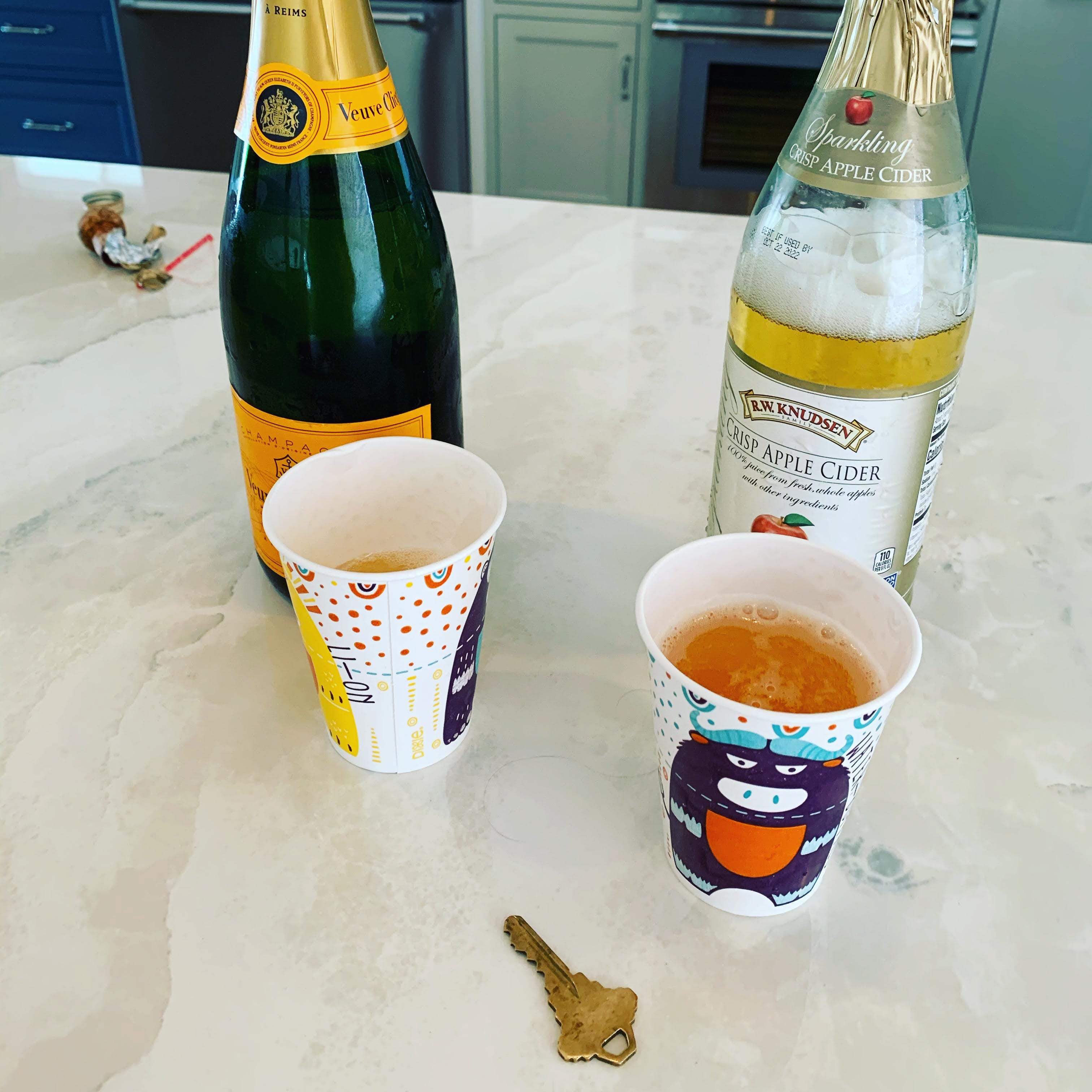 A bottle of champaign, a bottle of sparkling apple cider, two paper cups, and a house key.