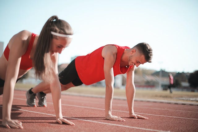 A man and a woman at a running track are beside each other in push-up position. They are wearing matching orange coloured athletic clothing maybe they are related? Who knows? My concern is if their push-ups are equal and the same.