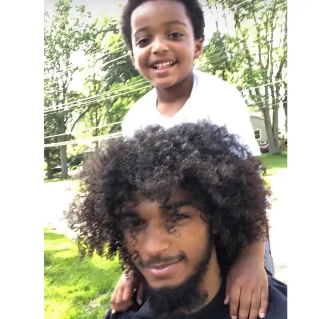 A photo of Casey Goodson, Jr. with a young boy smiling joyfully while riding on Casey’s shoulders. Greenery and a road are behind them. Casey is Smiling and camera-facing, with his hair longish and curly in front of his eyes.