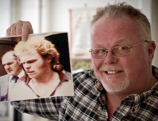 Now sporting white hair, a beard and moustache, and a receding hairline, Kirk Bloodsworth is shown holding up an older photograph of himself from the time of the initial trial. In the old photo, he has curly, reddish hair and bushy eyebrows.