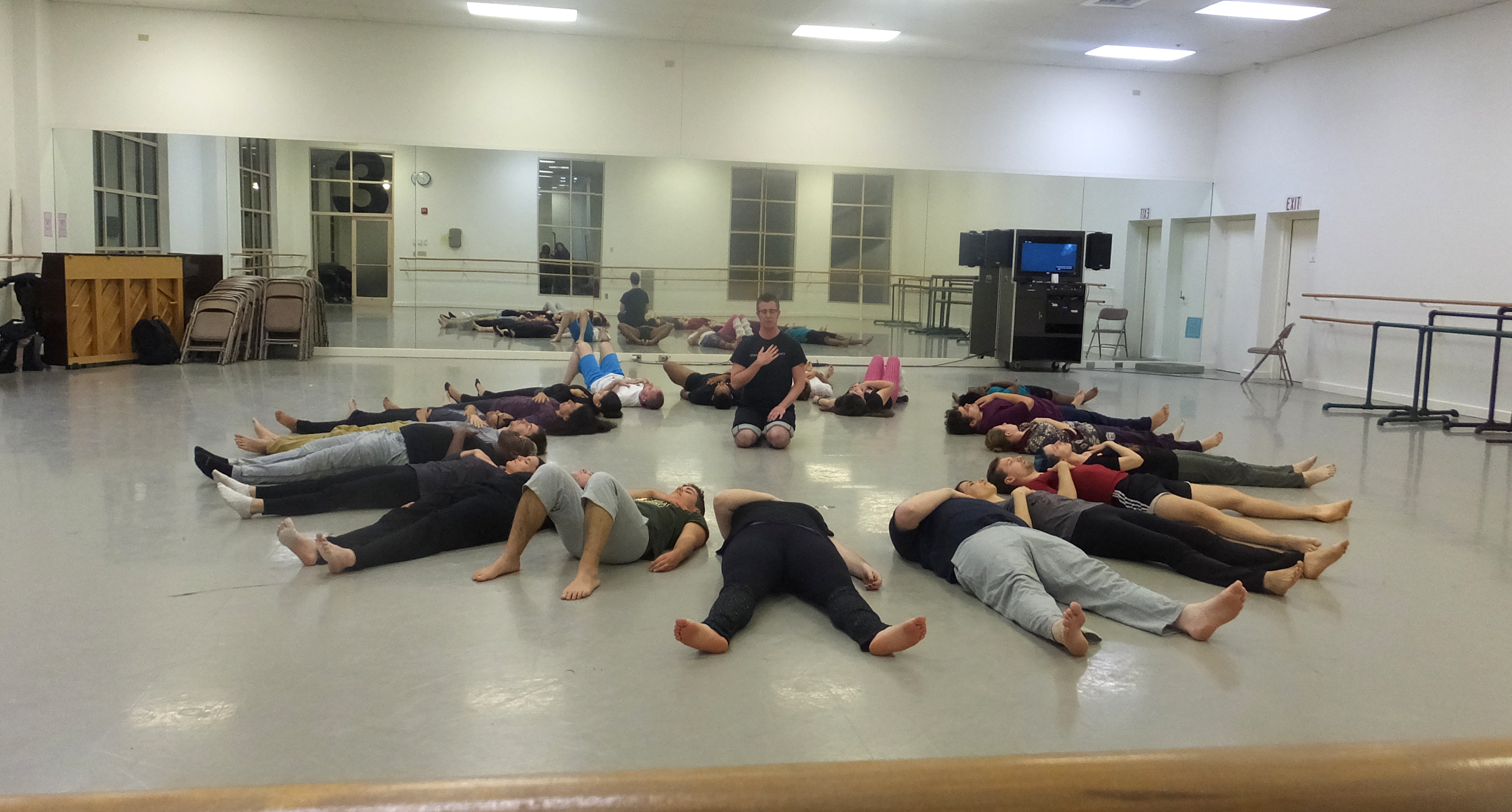 Group of people laying on their backs on the floor in a circle. Dorsey sits on his knees in the center with hand on heart.