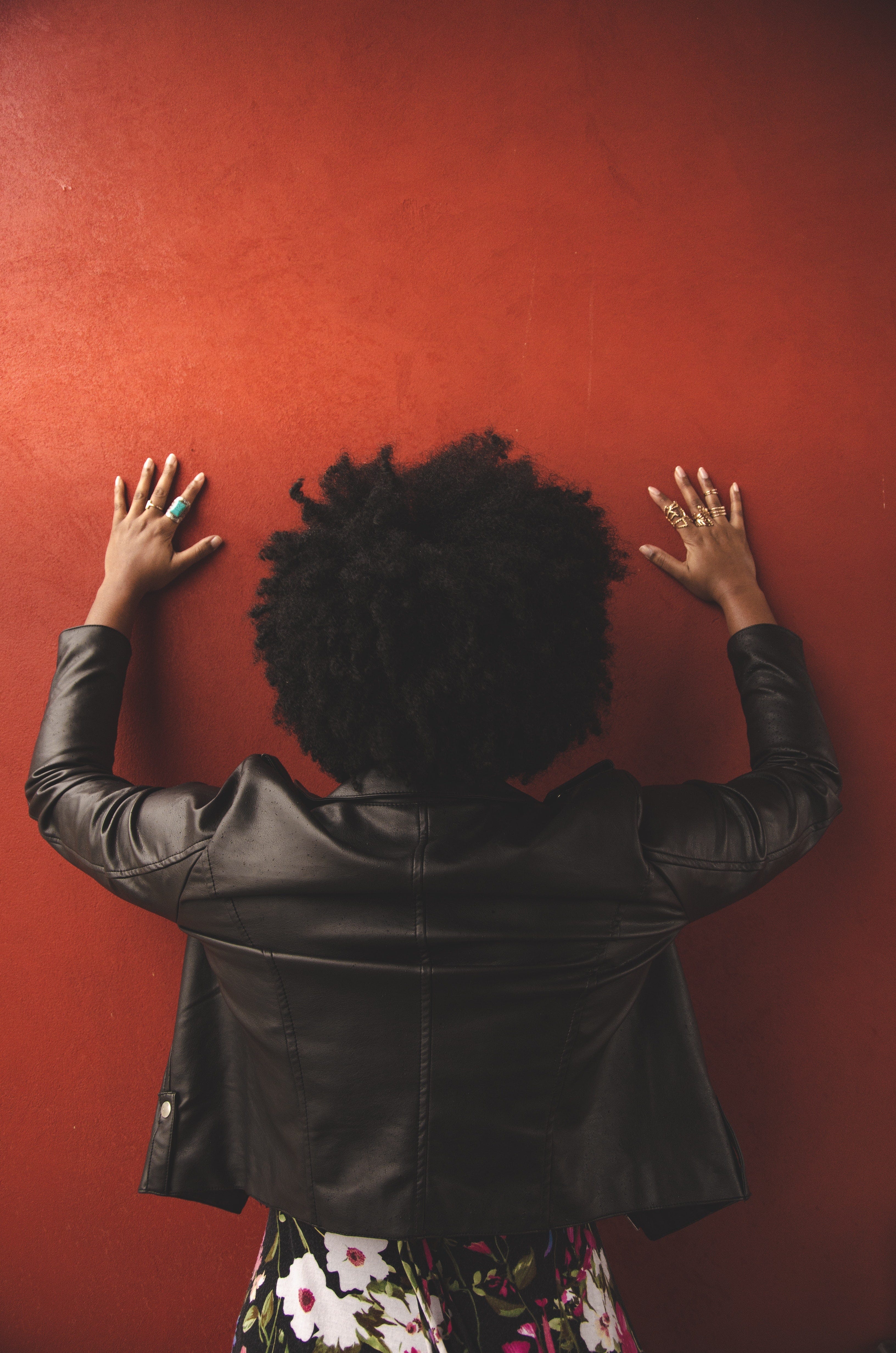 Image of a woman with afro hair taken from behind with hands up against the wall.