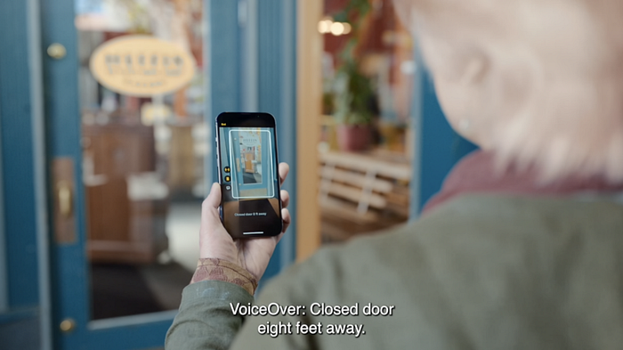 An elderly lady using Door Detection, holding her phone up to the door and the caption says: “VoiceOver: Closed door eight feet away”.