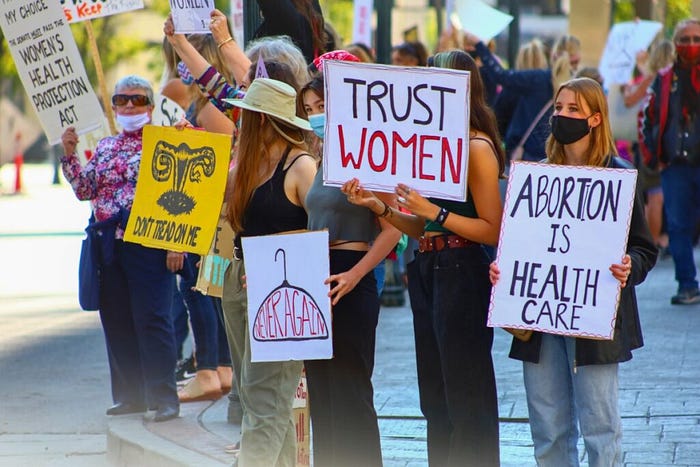 Photo of a protest about abortion rights. Signs read “Trust Women” and “Abortion is Health Care” and “Don’t Tread on Me.” One sign depicts a clothes hangar with the words “never again” inside.