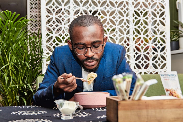 A person wearing a blue jacket, sitting at a table, eating a bowl of pasta or noodles in a pink bowl. There are plants in the backgrounn