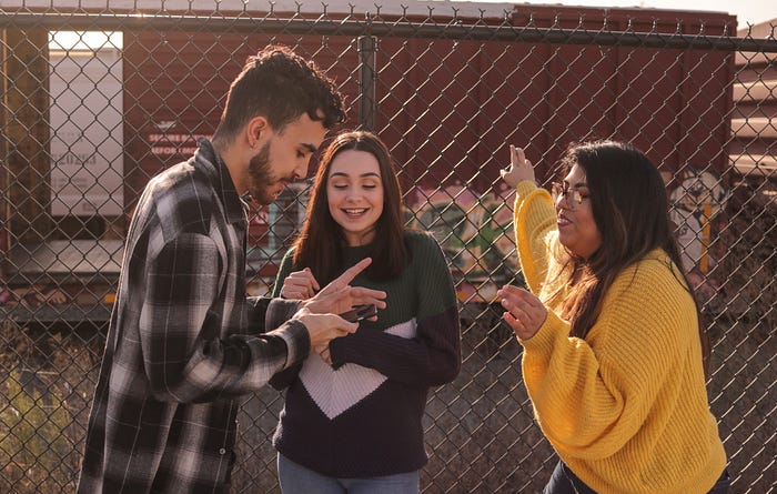 Three young people stand in front of a chainlink fence. Left is a young man with a beard, wearing a plaid shirt and holding a smartphone. Middle is a young woman with long brown hair, wearing a dark shirt, and right is another young woman with long dark hair, wearing glasses and a mustard yellow sweatshirt. They are all looking at the smartphone and smiling.