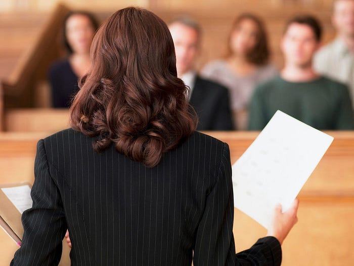 The back of a woman’s head in a courtroom featuring a blurred jury in the background.