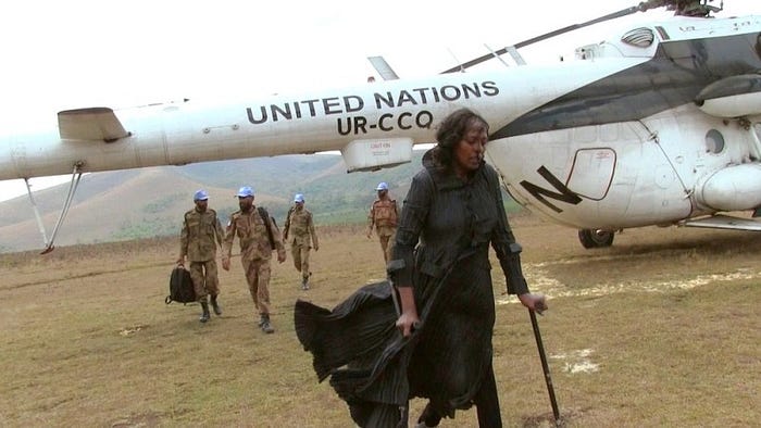 A picture of Neema Namadamu walking with crutches. Behind her, a UN plane and 4 men in military clothing.