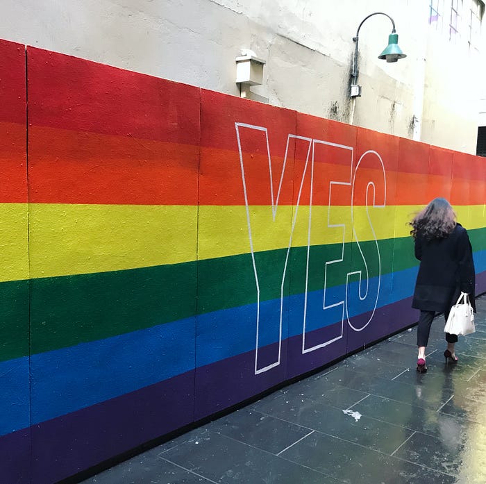 woman from behind walking in the street along a rainbow pride painted wall, with YES written on it