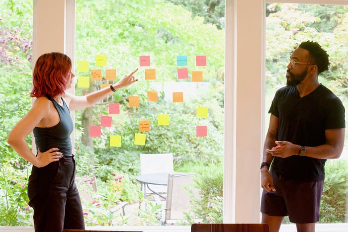 Two people standing in front of a window with sticky notes on it, discussing the data that they collected.