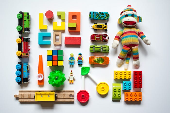 A set of colorful toys, including train cars, blocks, and hot wheels cars, is carefully arranged linearly in vertical and horizontal lines. There is also a sock monkey in a rainbow outfit.
