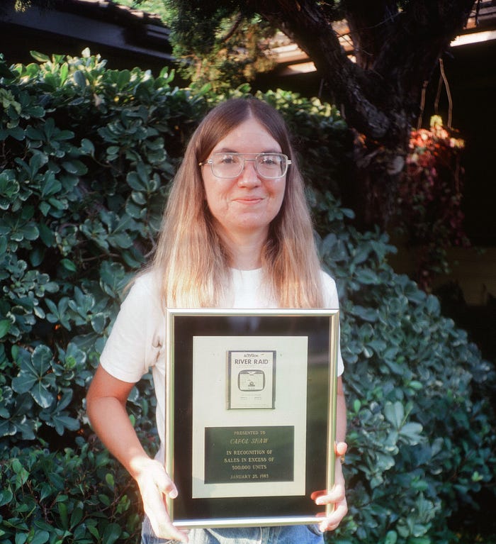 An older photograph of a young woman with long, straight, blonde hair and large, metal-phrame glasses. She wears a white shirt and jeans, is holding a silver award plaque, and stands in front of a green bush.