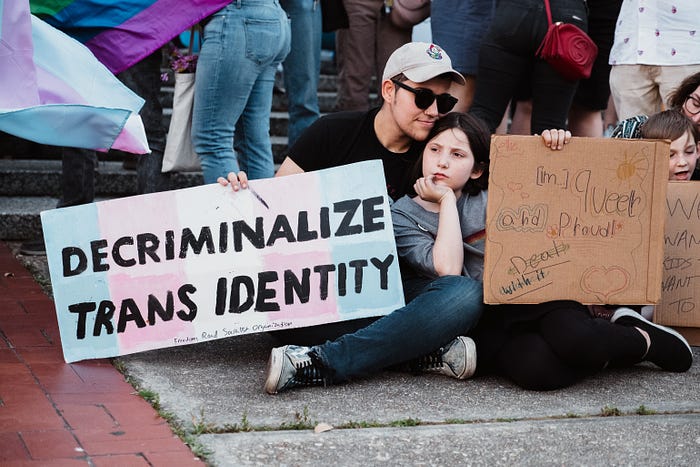 two people sit on a sidewalk with what looks like other people and pride flags in the background. The two people are an adult and a child. The adult holds a sign which reads: decriminalize trans identity. The child holds a sign that reads: I am queer and proud.