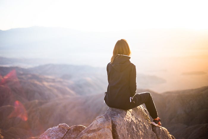 A woman sitting on the peak of a mountain facing away into the sunset.
