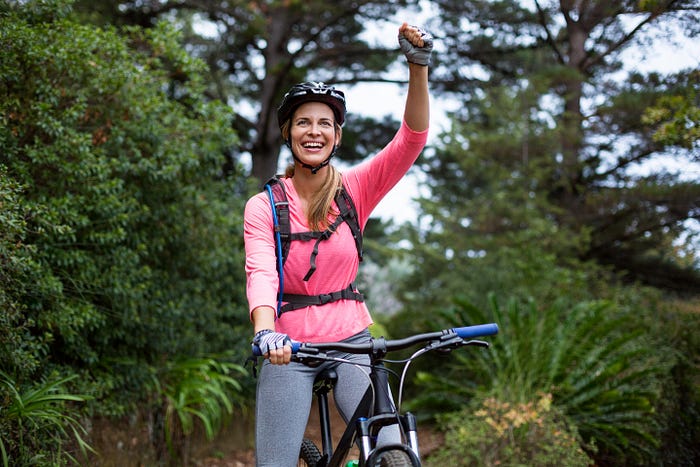 A woman cyclist raises her fist, she is wearing a hydration backpack while in the woods. Hydration Is Key, Stay Hydrated, Hydration Pack Essential