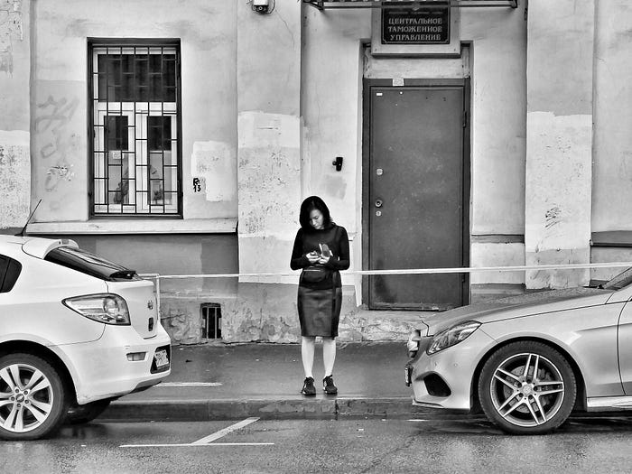 Black and white photo of woman in a black pencil skirt and black sweater, standing on sidewalk and looking at her photo, flanked by two cars parked on the street