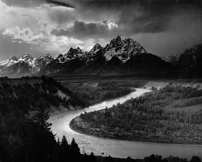 A black and white photo by Ansel Adams showing a winding river flanked by pine trees and leading toward the Teton mountains.