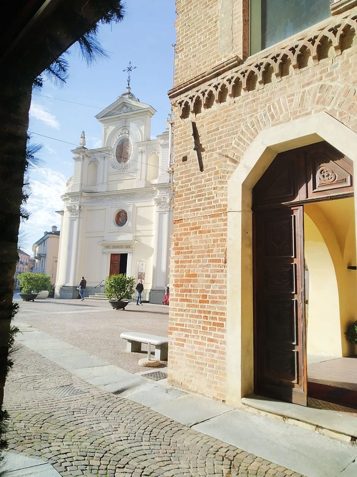 Italian plaza in alba, italia taken from a coffee shop called caffe vergnano. White historic church, sunny day, brick building.