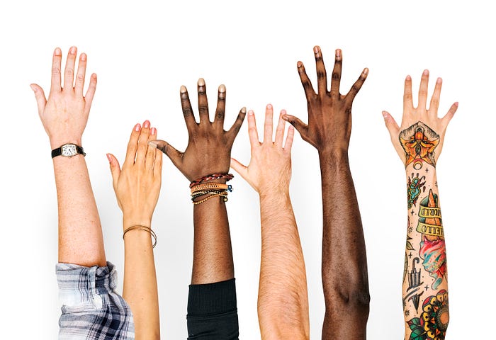 Six hands raised in the air. Physical characteristics indicate they are of different ethnicities, genders and subcultures.