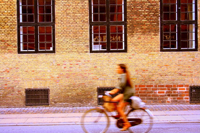 Woman in shorts on cycle, blurred, riding past old yellow brick urban factory building, in focus