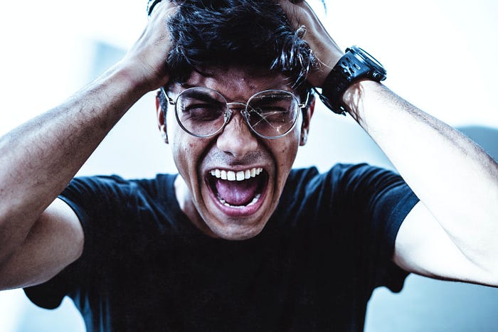 Man with short, dark hair, a black shirt and glasses holds his head while screaming in frustration.