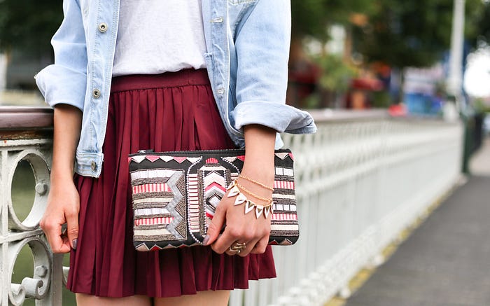 Middle of a person in a short, pleated, pocketless skirt, wearing bracelets and rings, and holding a strapless clutch purse.