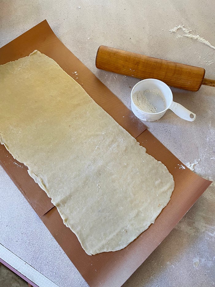 Pastry dough rolled out on a wax mat surrounded by a floured counter with a rolling pin and a cup of flour.