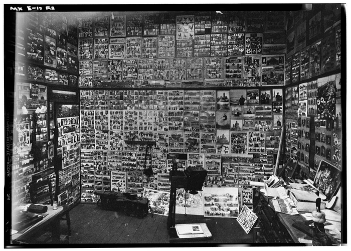 A black-and-white photograph of a photograph studio. Many pictures are plastered on the wall, occupying every visible surface.