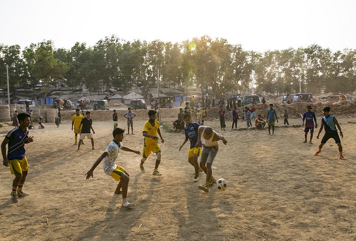 A group of boys plays football (soccer) on a dirt pitch.