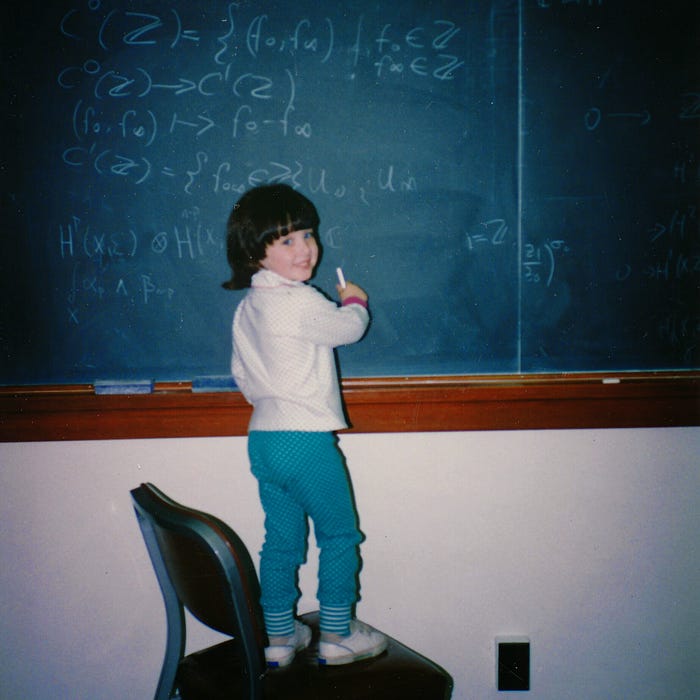 A little girl is standing on a chair at a blackboard. She has a white turtleneck shirt on and teal patterned pants. She’s holding a piece of chalk at the board and there is a math equasion after.