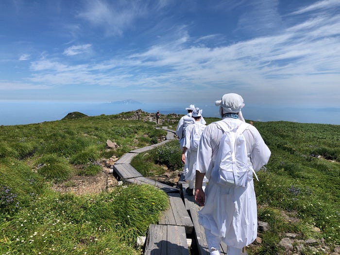 Yamabushi mountain ascetics walking on wooden planks on the path up Mt. Gassan of the Dewa Sanzan with Mt. Chokai in the background