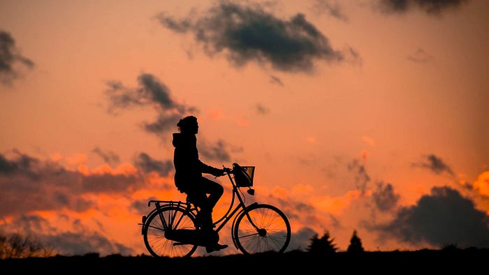 Photo of a silhouette of a woman on a bicycle, against an orange-colored sky at dusk with clouds