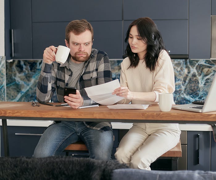 Two people sitting at a table. A man is holding a coffee mug but looking shocked at a piece of paper that the woman is holding, almost caught unawares by the news.