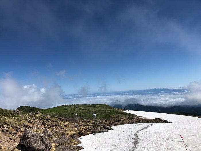 The path up Mt. Gassan of the Dewa Sanzan is covered in snow with clouds and the blue sky in the background.