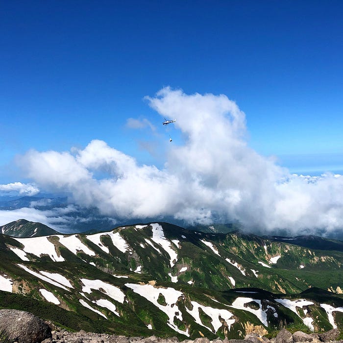 A helicopter flies through the clouds carrying a load in the sky above a snowy Mt. Gassan of the Dewa Sanzan.