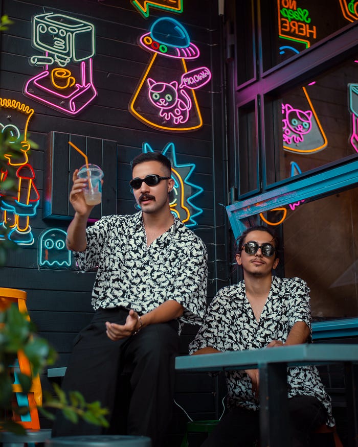 Two people with patterned shirts and sunglasses sit in front of a wall with many neon signs. The image gives the impression that the two people are cool and collected.