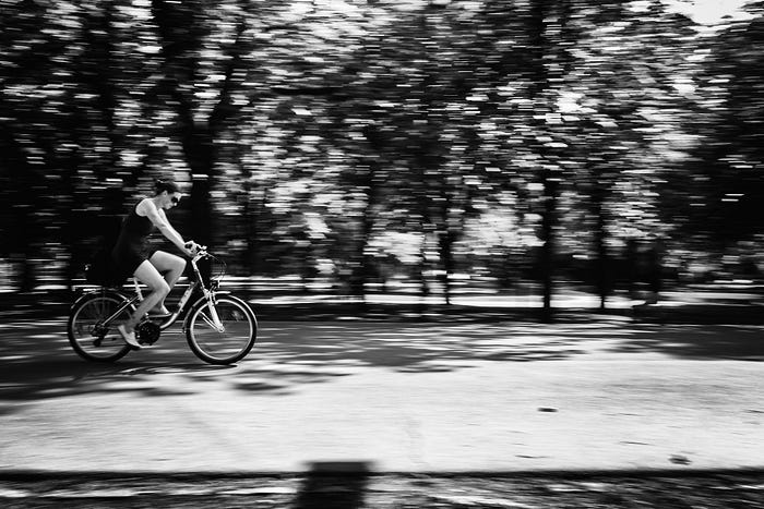 Black and white photo of a woman cycling past trees, with a slight motion blur